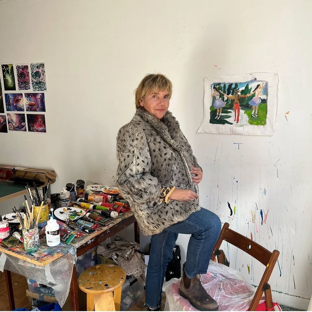 Delphine Perlstein assise au bord de sa table de travail dans son atelier, palettes et pinceaux à gauche, une toile accrochée au mur derrière elle.